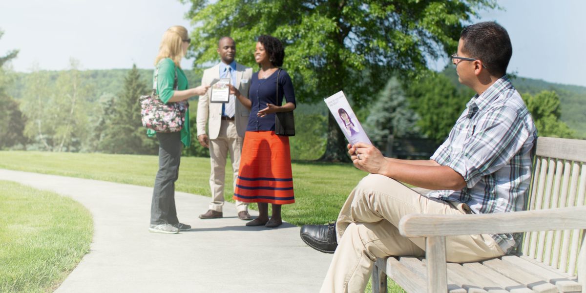 A man holds a tract and observes Jehovah’s Witnesses talking to a woman