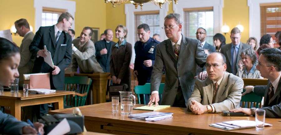 En la sala de un tribunal, los presentes reaccionando tras la lectura del veredicto. Un testigo de Jehová, sus abogados y varios hermanos están serios y tristes. Un grupo de no Testigos están sonriendo y felicitando a los abogados que ganaron el caso contra el hermano.