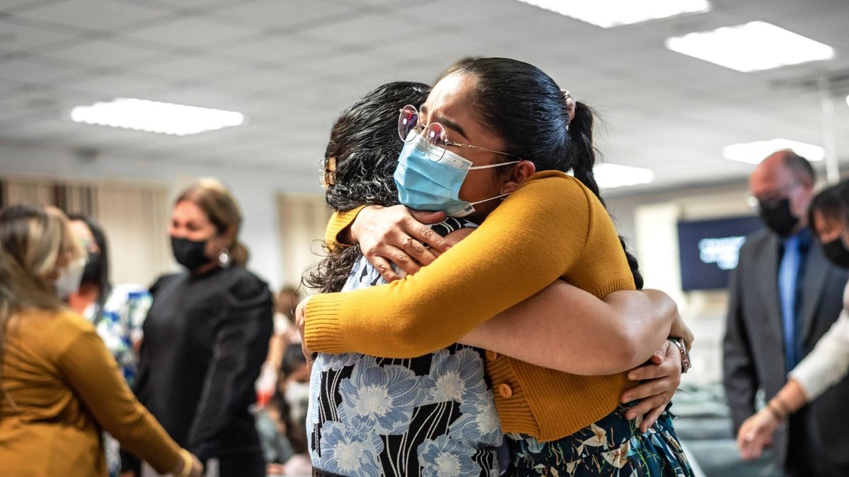 Dos hermanas se abrazan felices en el Salón del Reino. Todos los asistentes llevan puesta la mascarilla o cubrebocas.