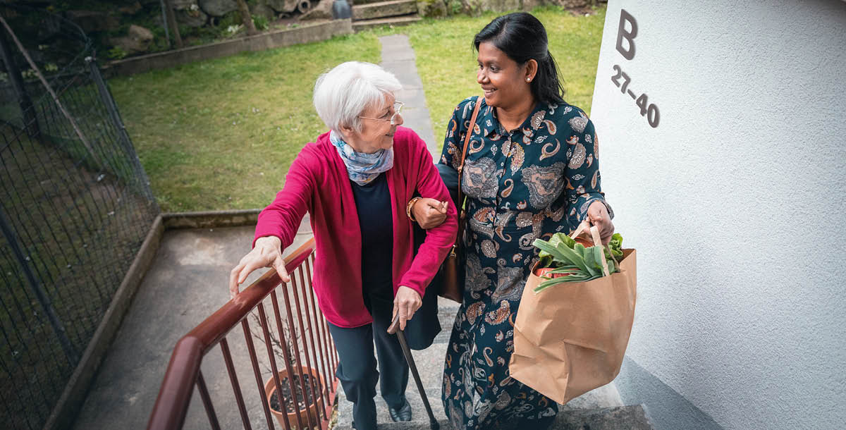 ’n Indiese vrou help ’n ouer Kaukasiese vrou met haar inkopies by die trappe op.