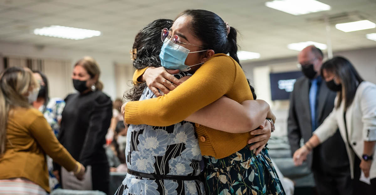 Twee susters dra maskers en gee mekaar ’n drukkie by ’n Koninkryksaal. In die agtergrond dra broers en susters maskers terwyl hulle mekaar liefdevol groet.
