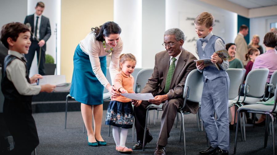 ’n Ouerman gesels met jong kinders by die Koninkryksaal en kyk na prentjies wat hulle geteken het.