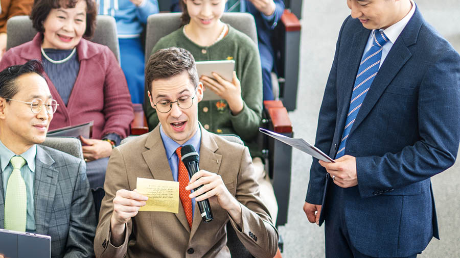 By ’n vergadering by die Koninkryksaal lees ’n broer wat ’n ander taal leer, sy antwoord af wat hy op ’n papier neergeskryf het. Die ander wat in die saal sit, kyk met respek na hom.