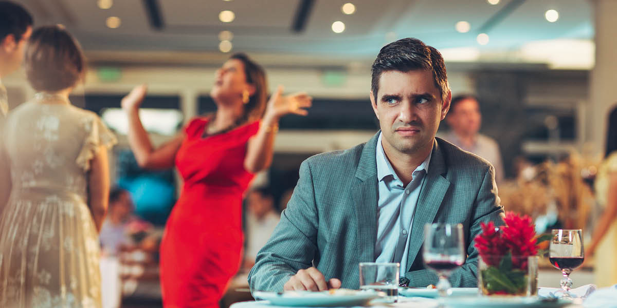 رجل مزعوج يجلس وحده على طاولة في مطعم بينما تستمتع زوجته بالحديث مع زوجين