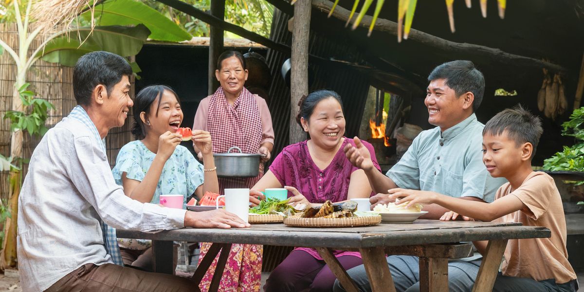 A family gathered around a table outside their modest home, happily sharing a meal with a guest.