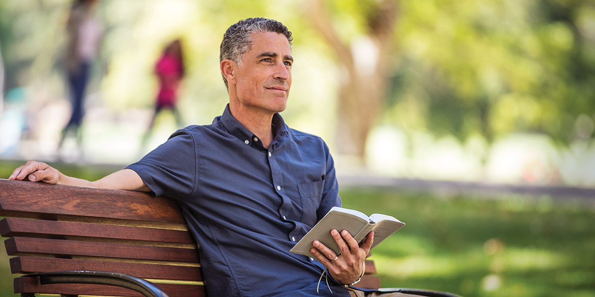 A man who is coping with suicidal thoughts sits on park bench, holds an open Bible, and meditates