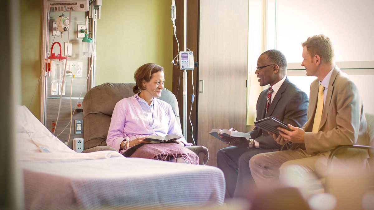 Two elders visiting a sister in the hospital. Their Bibles are open as they speak with her.