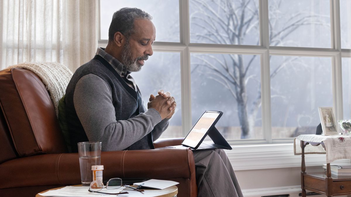 An elderly brother sitting in his home and praying in the wintertime. An open Bible is on his lap, and a bottle of medication is on a table next to him.