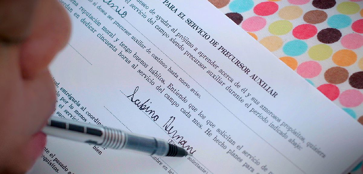 Si Sabina Hernández na igwang bolpen sa saiyang nguso asin pinipirmahan an aplikasyon para sa pag-auxiliary payunir