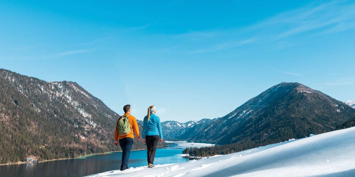 Um casal caminhando em uma encosta coberta por neve. Eles observam um lago que é cercado por montanhas e árvores, debaixo de um céu azul.