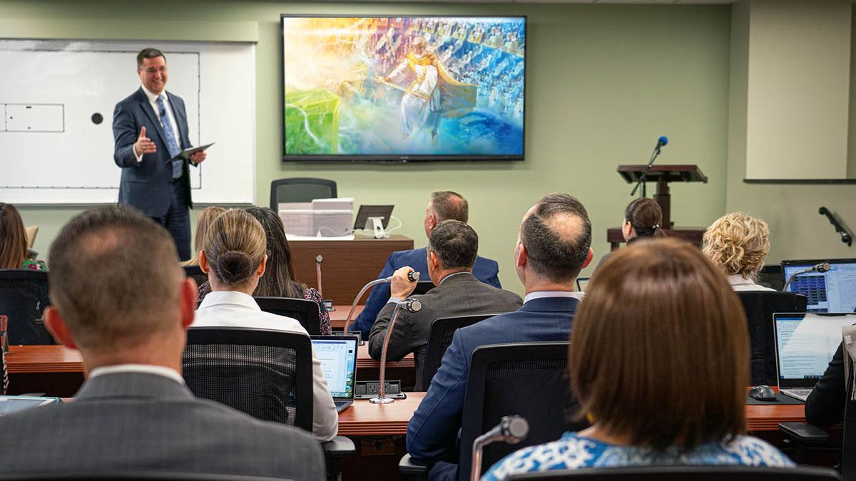 Um instrutor dando uma aula. Atrás dele, uma TV está mostrando uma imagem de Jesus entregando o Reino para Jeová. Os alunos usam os microfones que estão nas mesas para comentar.