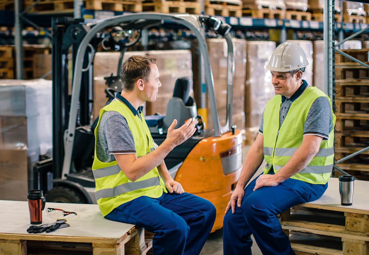 Um irmão sentado numa mesa dando um convite da Celebração para um parente que não é Testemunha de Jeová enquanto as esposas conversam e preparam uma refeição juntas.
