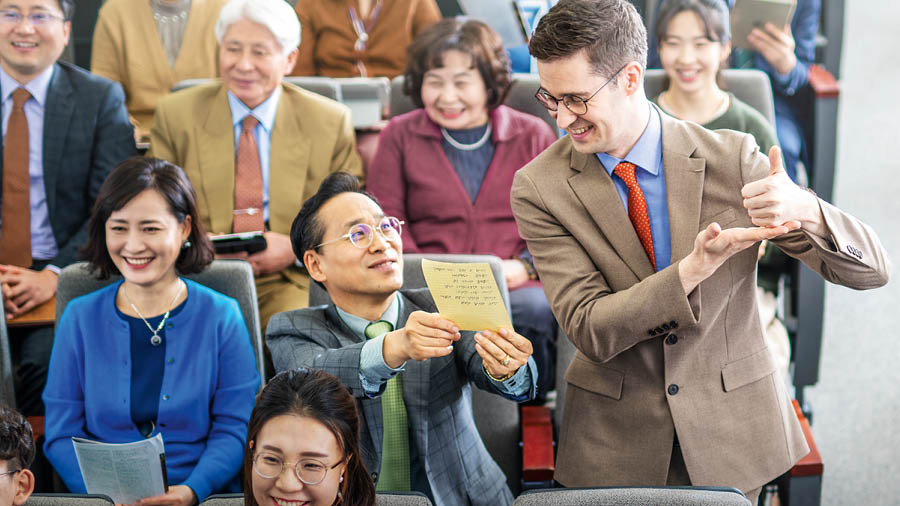 Durante uma reunião, um irmão que está aprendendo um idioma lê um comentário que ele escreveu à mão. Os outros irmãos na assistência olham com respeito.
