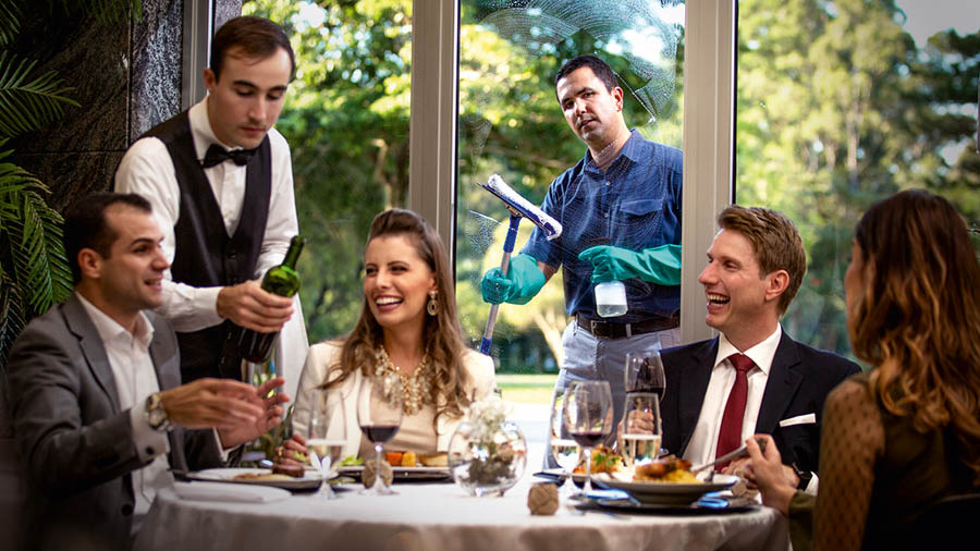 Dois casais rindo e aproveitando uma refeição num restaurante caro. O irmão que está limpando a janela olha para eles e sente inveja do seu aparente sucesso.