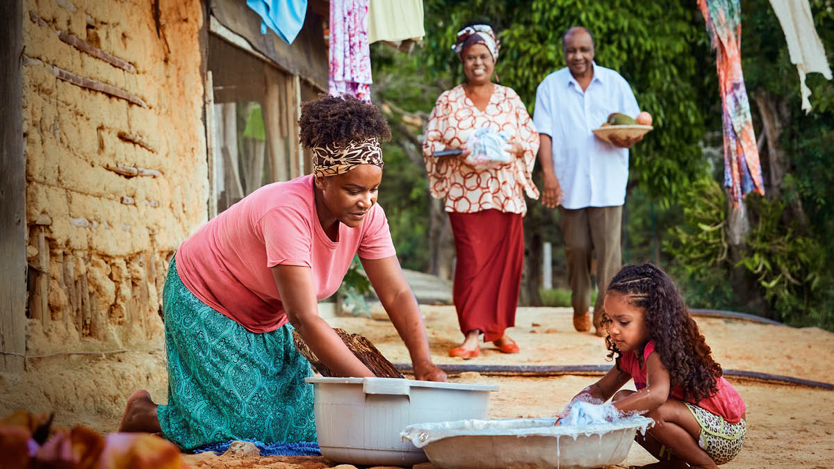 Uma irmã e sua filha pequena lavando roupas à mão. Um casal está trazendo comida para elas.