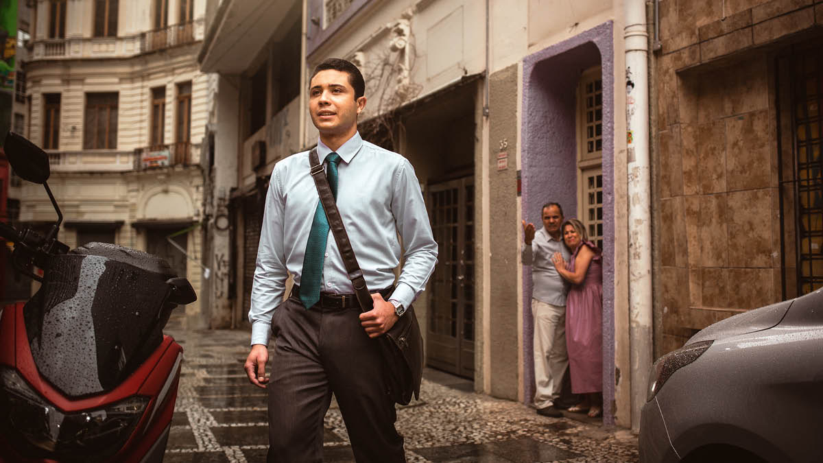 Um irmão jovem vestido para ir a uma reunião. Ele está andando confiantemente. Ao fundo, seus pais estão na porta da casa gritando com ele.