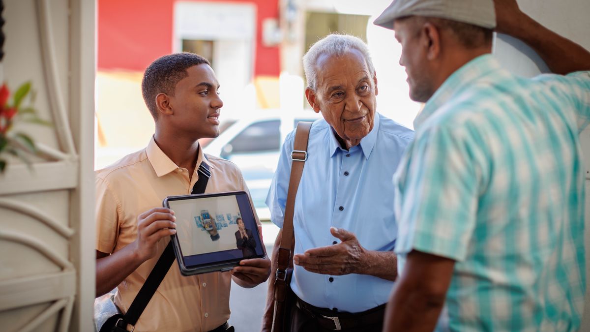 Um irmão jovem e um irmão idoso trabalhando juntos na pregação de casa em casa. O irmão idoso conversa com o morador enquanto o irmão jovem segura um tablet para mostrar um vídeo.