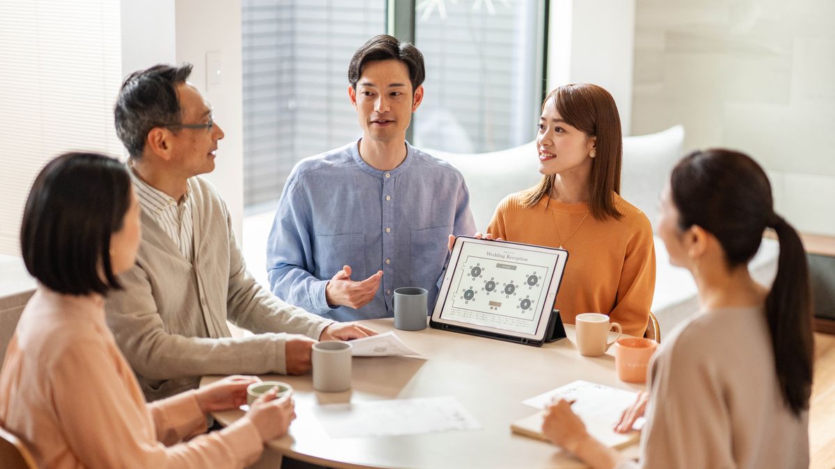 Um casal que está noivo conversa sobre os planos do casamento com alguns amigos que vão ajudá-los. O noivo segura um tablet que mostra as mesas dos convidados na festa de casamento.