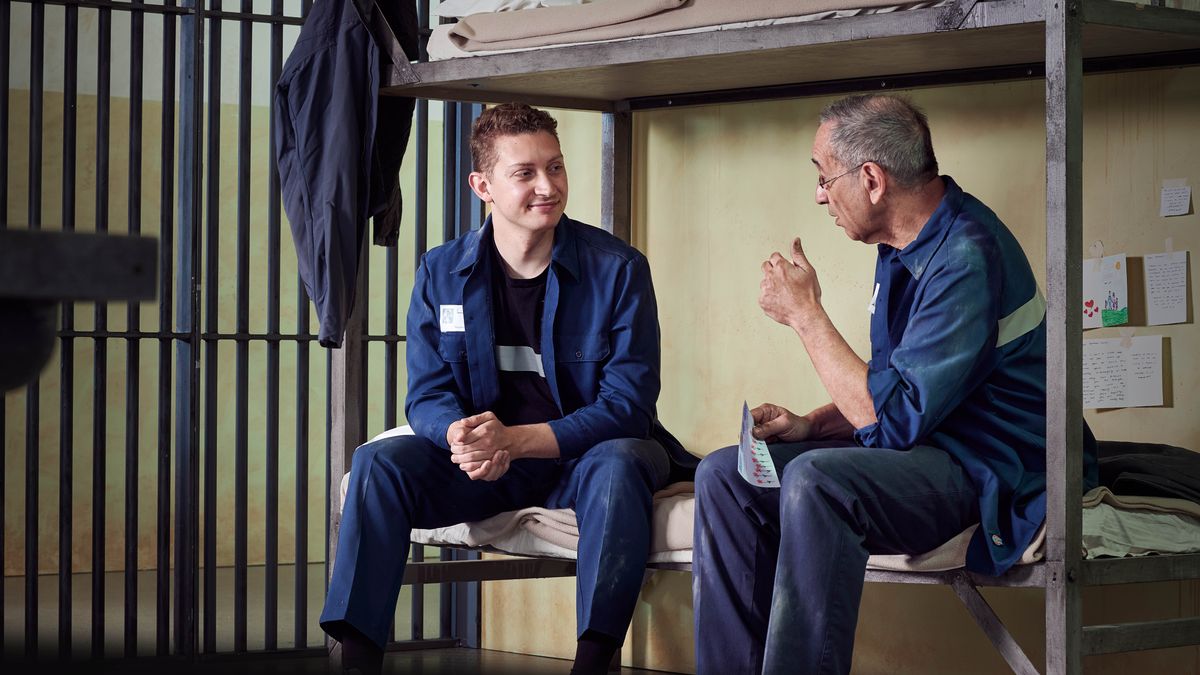 Dos germans conversant mentre seuen al llit de la cel·la d’una presó. A la paret darrere d’ells hi ha penjades postals, cartes i un dibuix.