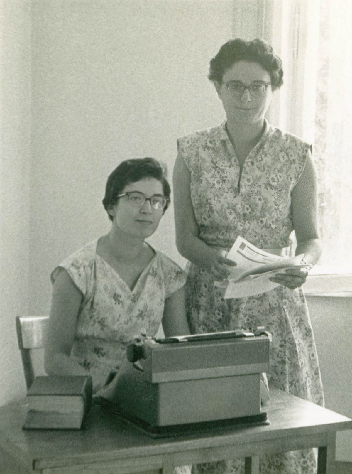 Si Irma naglingkod nga naay typewriter sa iyang atubangan, ug si Ilaria nagbarog tapad niya.