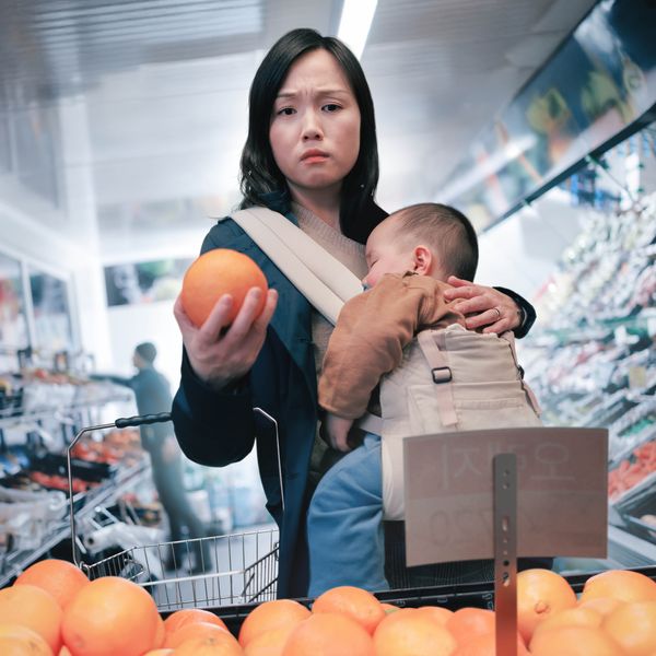 En un supermercado, una madre que lleva a su bebé dormido mira preocupada el precio de la fruta.