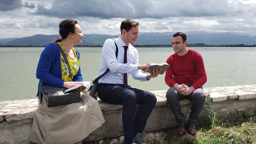 Alida og Stefano forkynder for en mand; de sidder alle på en stenmur ved en sø.