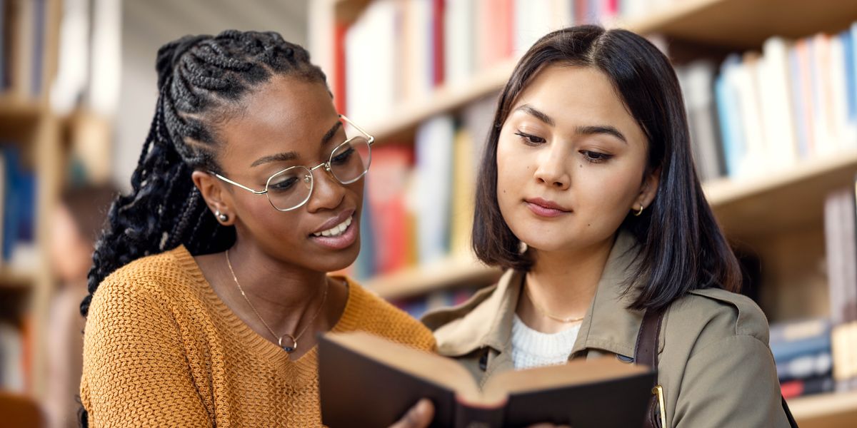 In einer Bibliothek schauen zwei junge Frauen in ein Buch.