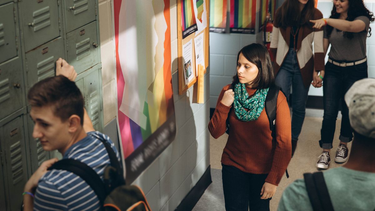 Die Schwester geht einen Schulflur entlang und schaut auf ein Poster, das für homosexuelle Lebensstile wirbt. Hinter ihr gehen Hand in Hand zwei Schülerinnen, die Armreifen in Regenbogenfarben tragen.