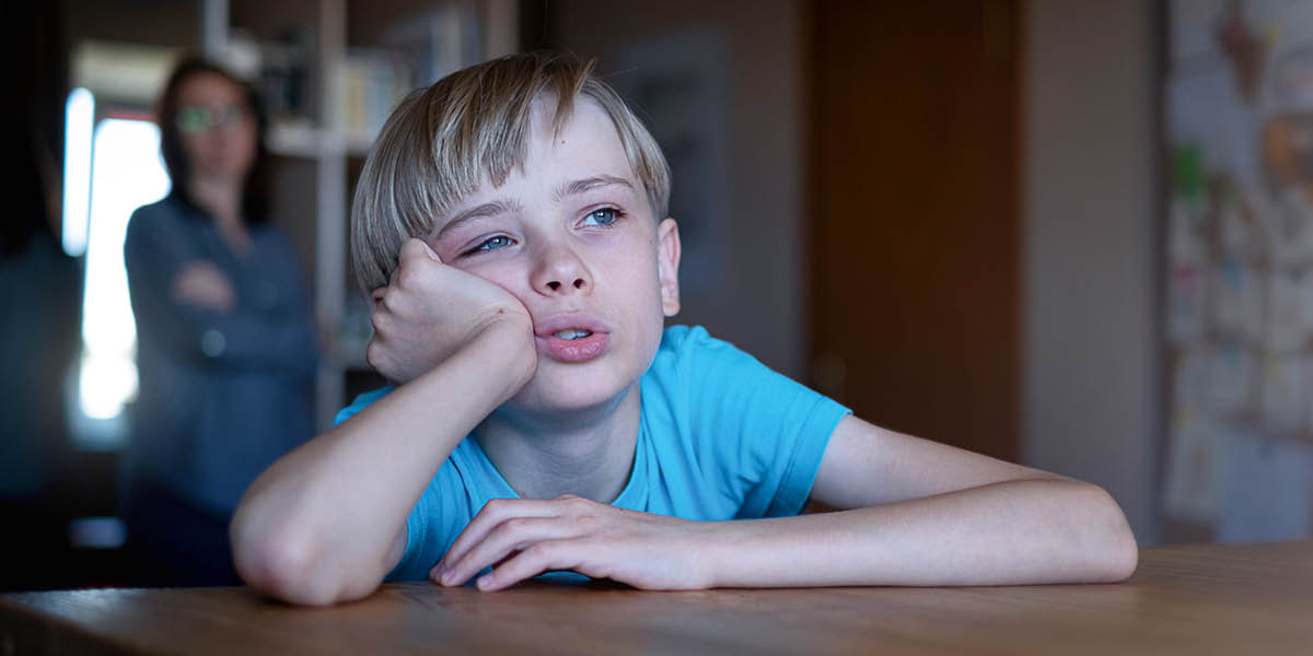 Un niño apoyado en una mesa mira por la ventana con cara de aburrimiento. Al fondo su mamá lo observa preocupada.