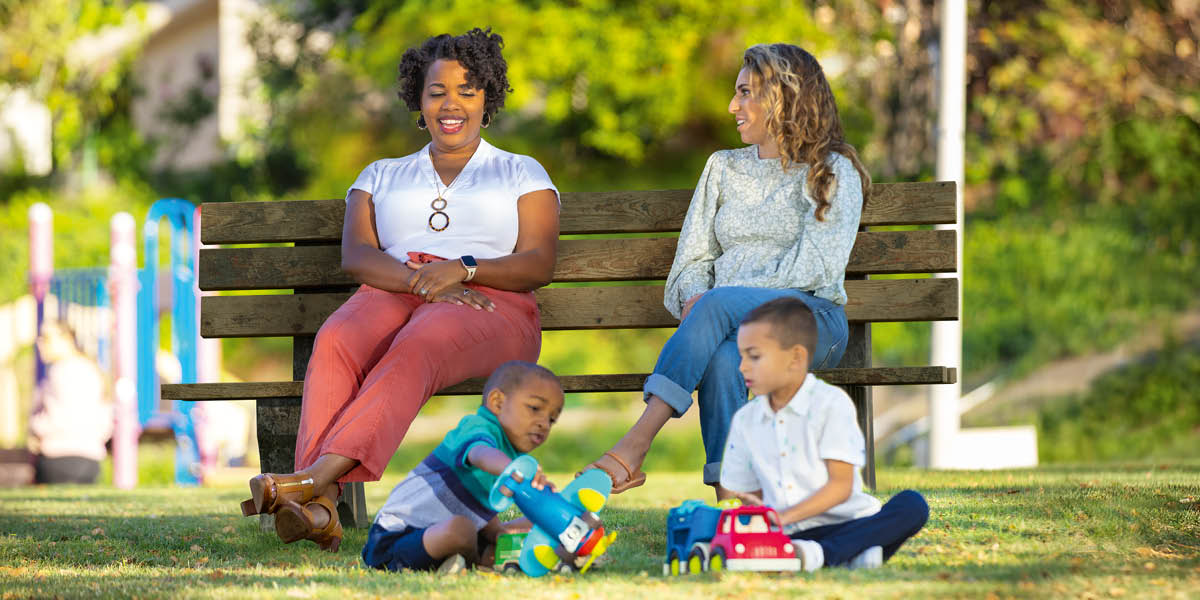 Two mothers of different races sitting on a park bench, smiling and talking while their two young sons play together.