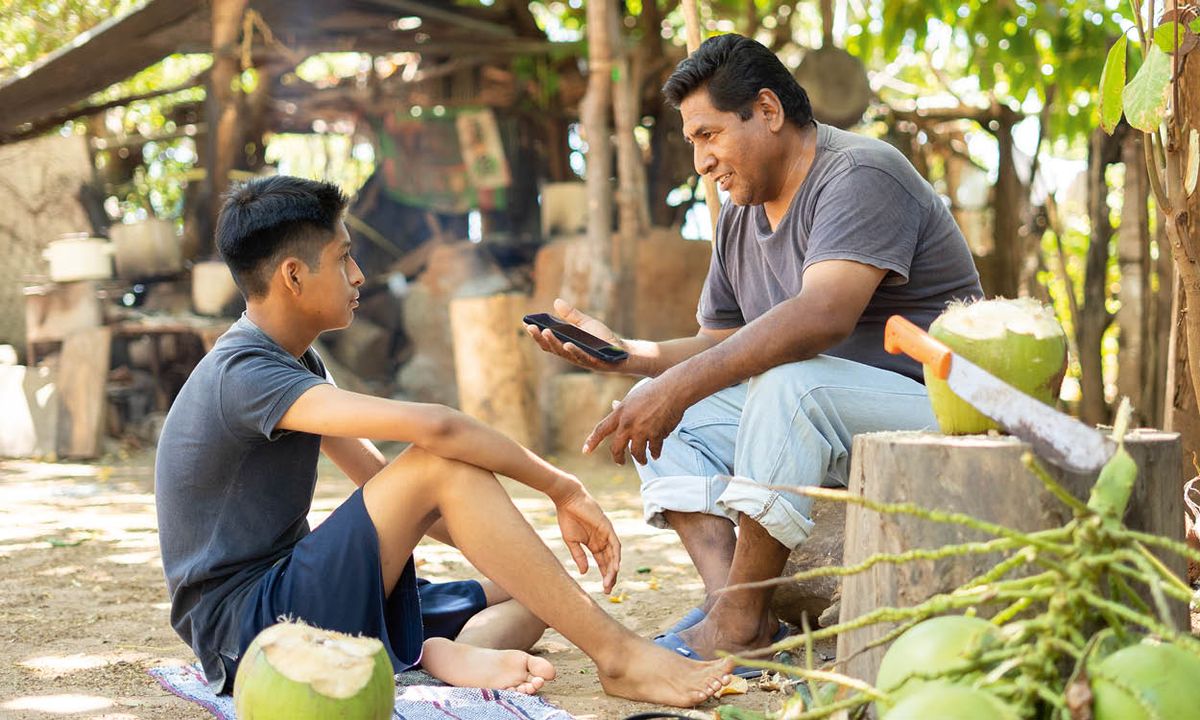 A father holding a cell phone and talking with his young son outdoors.