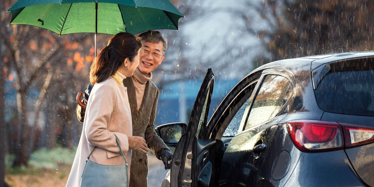 An older man holding an umbrella over his wife’s head as he opens the car door for her.