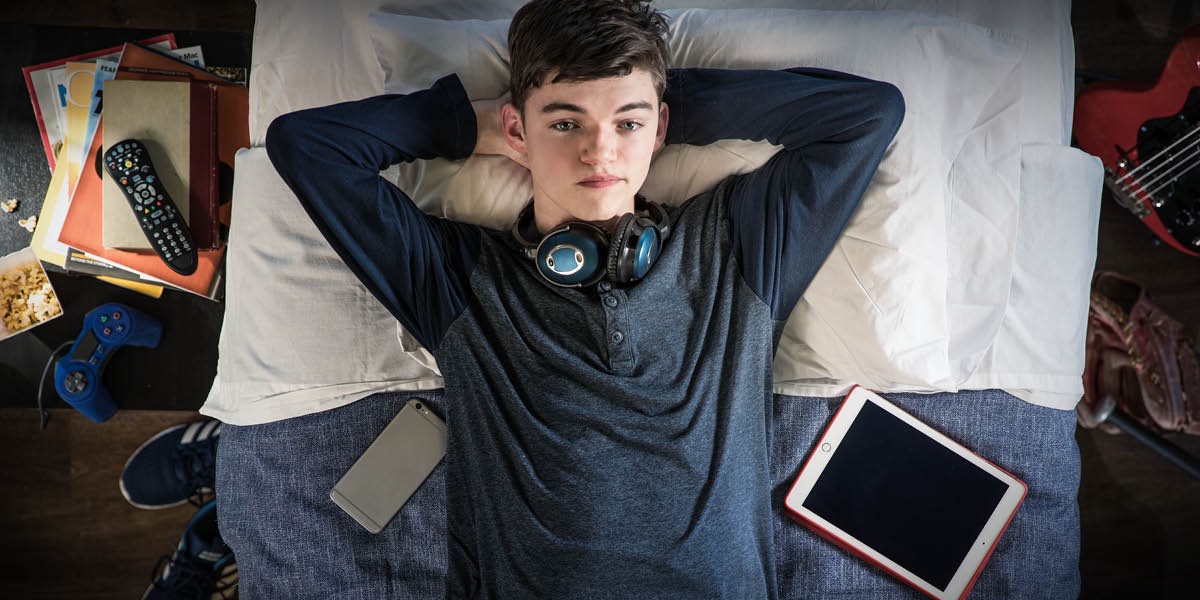 A teenage boy has many electronic devices, but he lays on his bed and stares at the ceiling