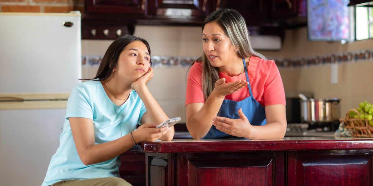 A frustrated teenage girl holding her phone while her mother tries to reason with her.