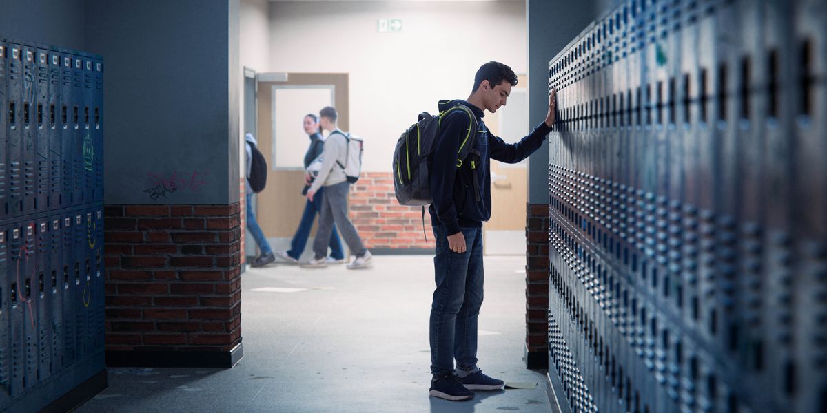 A downcast teenage boy standing by a row of lockers in a school hallway.