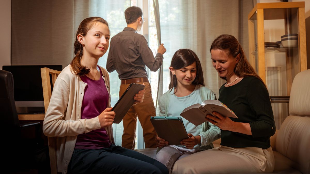 A family studying the Bible together in a land where our work is restricted. The father keeps watch at the window of their home while the mother reads the Bible with their two daughters.