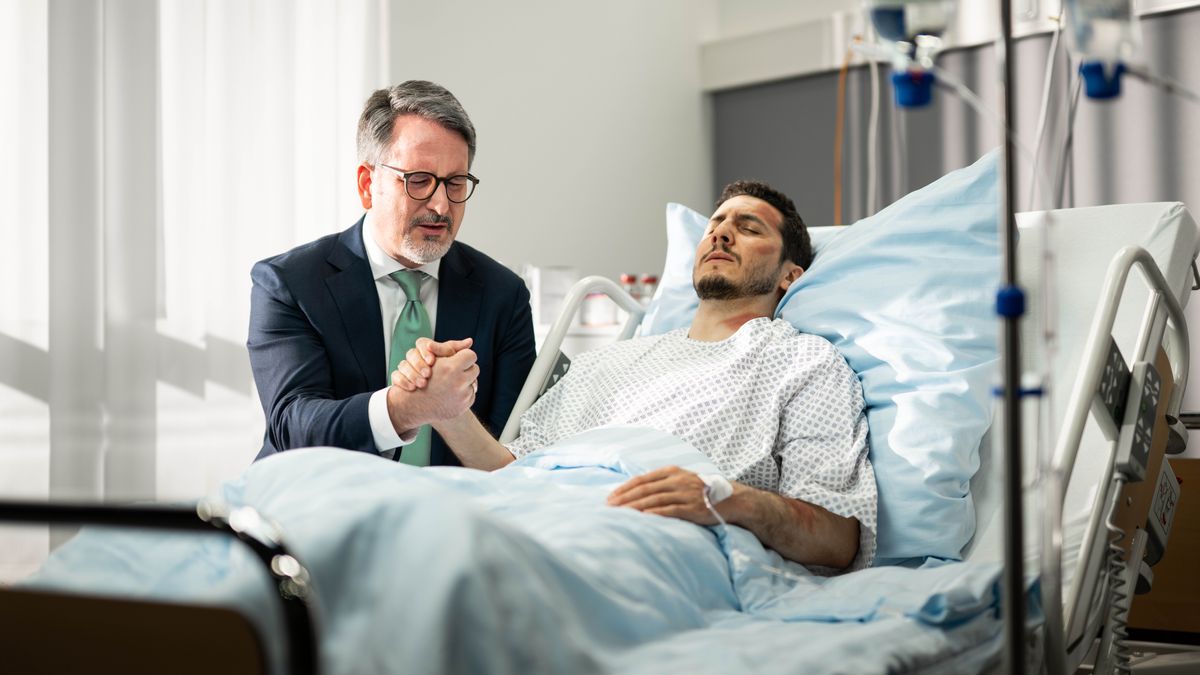 A brother prays at the bedside of a brother who is in the hospital and covered in bruises.