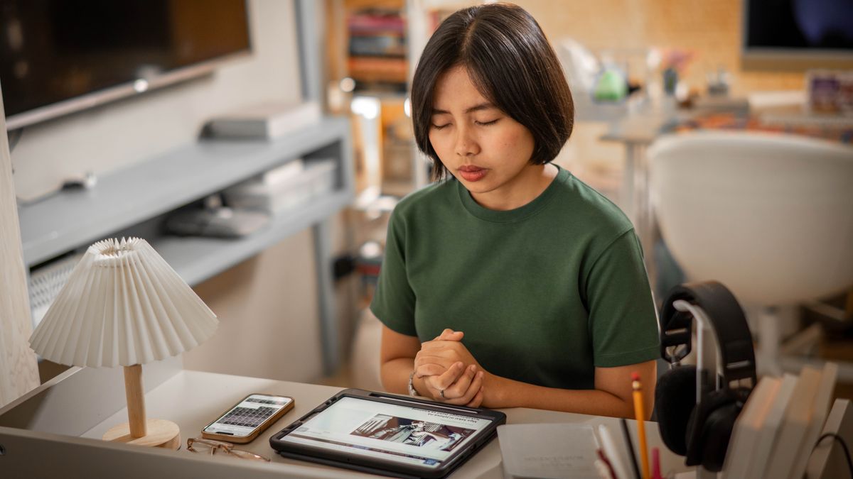 A young sister prays at her desk. Her tablet displays an article on jw.org about brothers and sisters who are imprisoned for their faith.