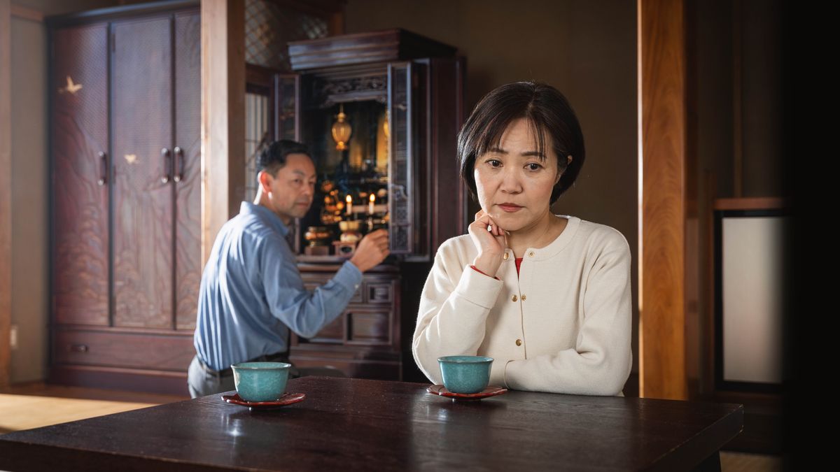 A sister thinking deeply while her husband kneels before a Buddhist altar at their home.