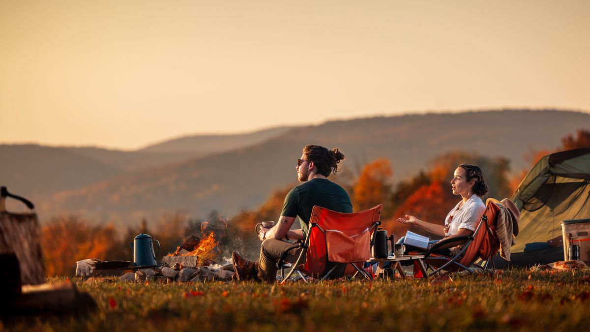 A sister speaking to her unbelieving husband while they enjoy some coffee near a campfire. She has her Bible open and gestures toward the beautiful scenery.