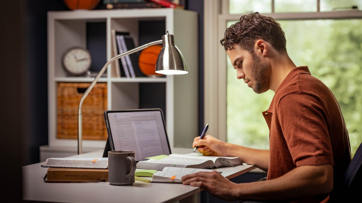 Un hermano joven está haciendo su estudio personal con la Biblia, una tableta y un cuaderno, y está anotando algo en la Biblia.