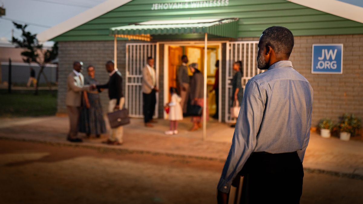 Un hermano mayor con la Biblia en la mano está de pie mirando un Salón del Reino que está a unos cuantos metros de distancia. Observa a los hermanos y a las hermanas saludándose con cariño.
