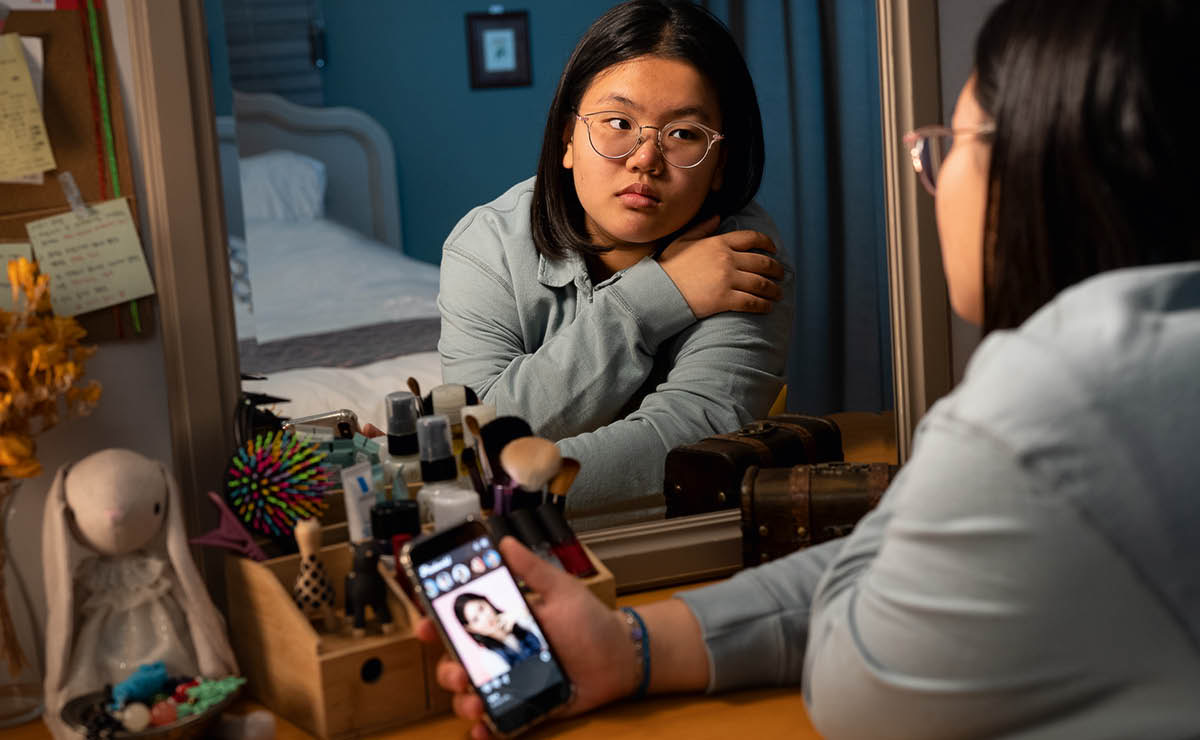 Une adolescente se regarde dans un miroir et se compare avec une fille dont la photo est affichée sur son téléphone.