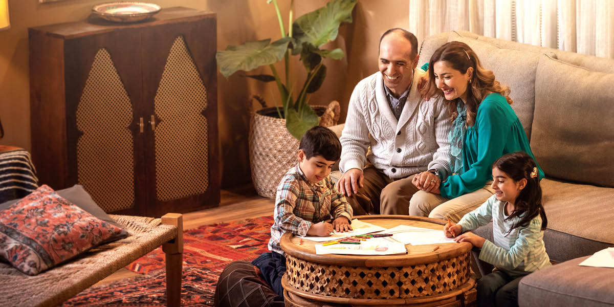 Une famille, heureuse de passer du temps ensemble. Les parents, assis sur le canapé, se tiennent la main. Les enfants, installés à la table basse, dessinent.