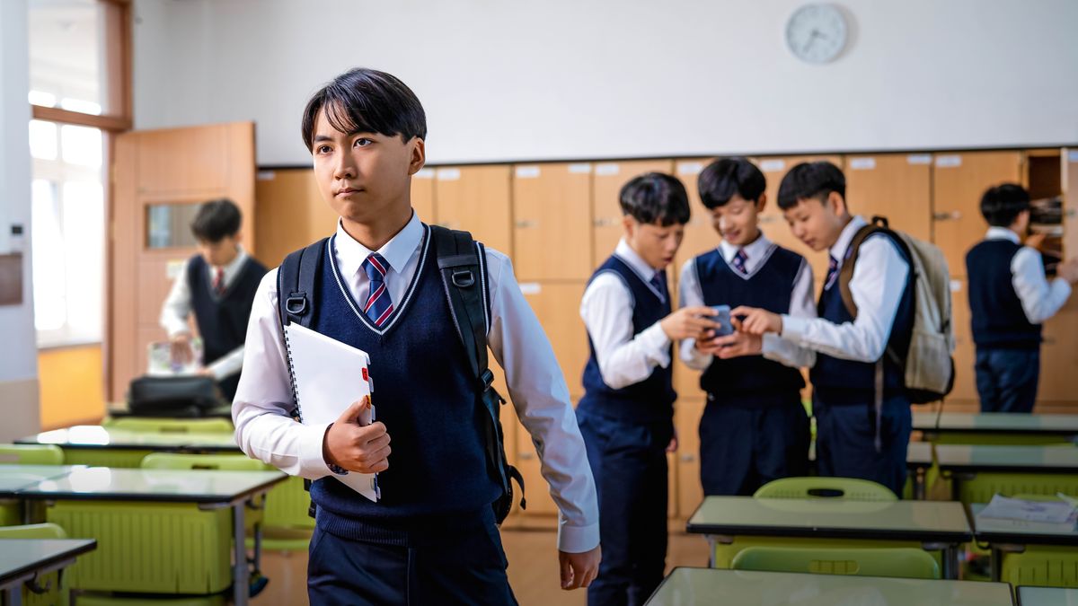 A young brother at school, confidently walking away from his classmates who are looking at something on a smartphone.