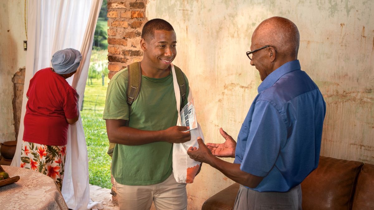 A brother entering the home of a Witness couple. He gives the husband a printed publication while the wife keeps watch at the entrance of the home.
