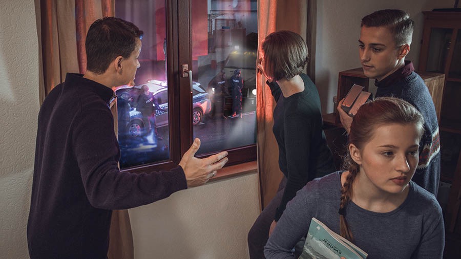 Dieselbe Familie. Die Eltern beobachten vom Fenster aus Polizisten vor ihrem Haus, während der Sohn und die Tochter ganz ruhig darangehen, biblische Veröffentlichungen zu verstecken.