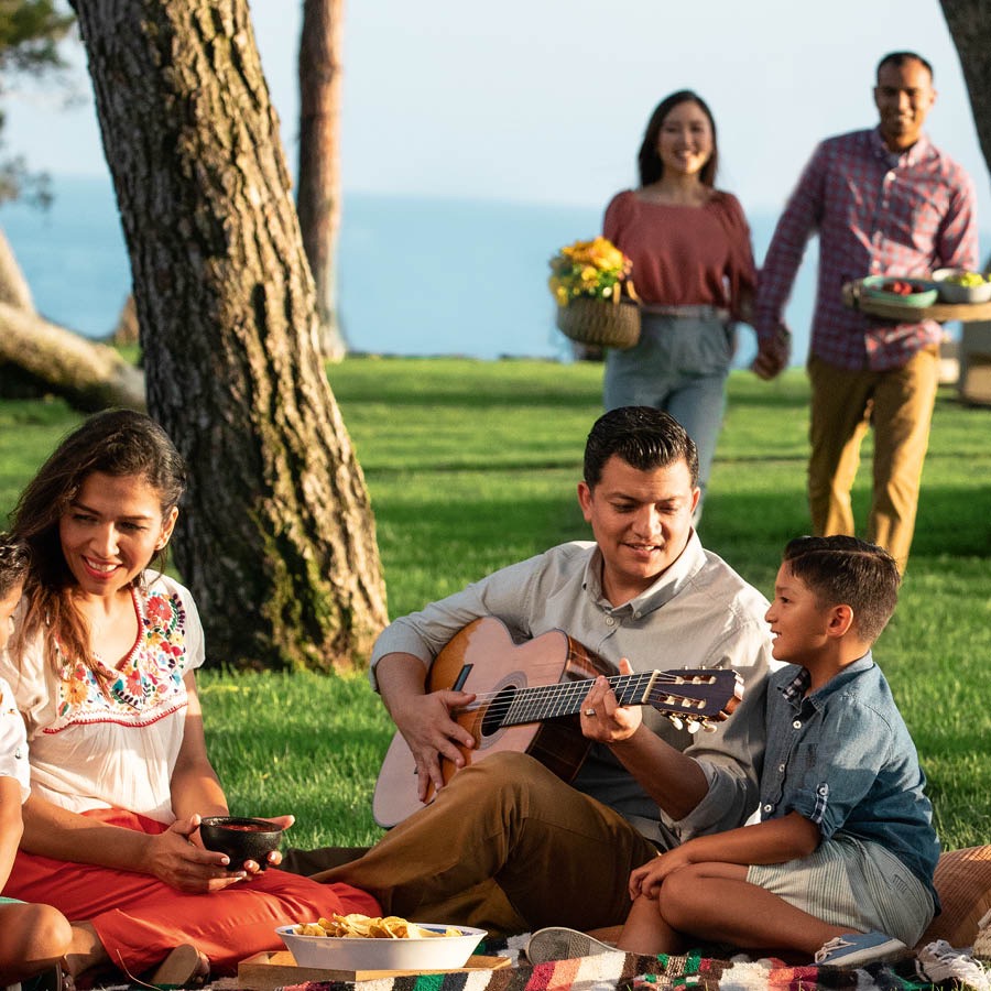 Eine Familie macht in einem Park ein Picknick. Der Vater spielt Gitarre.