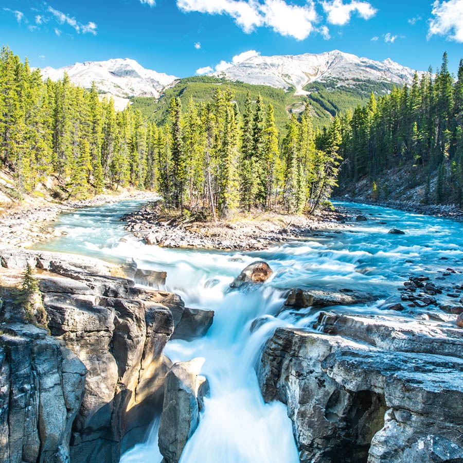 Ein Fluss geht in einen Wasserfall über. Im Hintergrund sieht man schneebedeckte Berge und einen Wald.