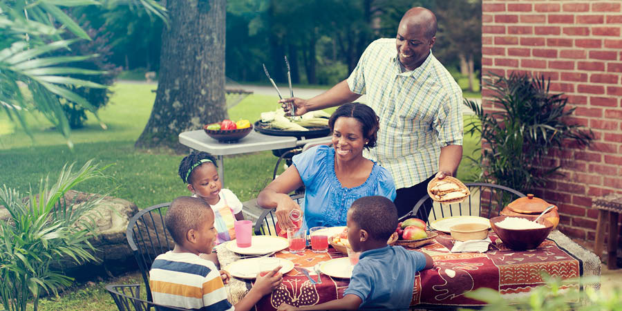 Eine glückliche Familie sitzt gemeinsam beim Essen im Garten. Der Vater grillt und bringt das Essen zum Tisch.